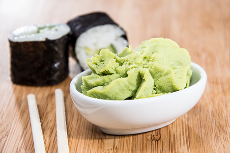 Wasabi preparado en un plato junto a dos palillos. Atrás hay dos rollos de sushi. Ambos alimentos están sobre una mesa de madera. 
