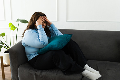 Una mujer con sobrepeso triste sentada en un sofá sufriendo los estigmas sociales asociados al peso. 