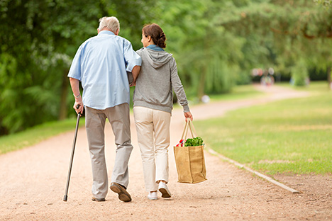 Hombre mayor es ayudado a andar por una mujer que carga las compras del anciano. 