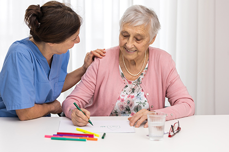 Una adulta mayor dibujando en compañía de una enfermera. 