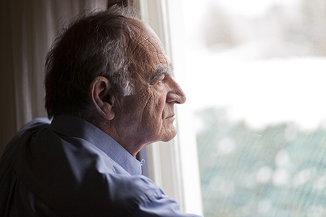 Un adulto mayor observando su entorno con tristeza desde una ventana. 